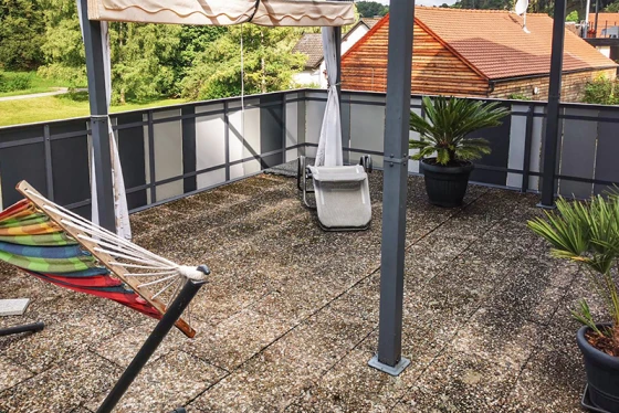 Strešna terasa s starimi betonskimi ploščicami ponuja prostorno bivalno površino. Umazana površina ploščic kaže očitne znake obrabe. Senčilo nudi prijetno senco v vročih dneh. Lonci s palmami in viseča mreža ustvarjajo prijetno vzdušje. Kovinska in steklena ograja dopolnjujeta sodoben in odprt dizajn terase.