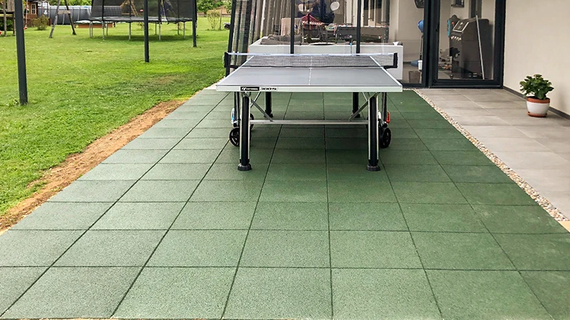Na terasi, obloženi z zelenimi WARCO ploščami iz PU vezanih gumijastih zrn, je pred razkošno hišo postavljena miza za namizni tenis. Na vrtu se nahajata trampolin in druga igrala, ki vabijo k gibanju in sprostitvi. Te robustne, nedrseče plošče zagotavljajo udobje in varnost. Njihova elastična površina varuje sklepe, medtem ko enostavno vzdrževanje ohranja urejen videz skozi daljše obdobje.