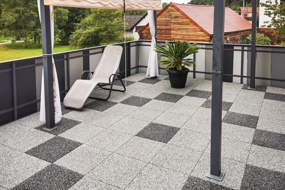 Obnovljena strešna terasa je opremljena z granitnimi ploščicami, razporejenimi v geometričnem vzorcu svetlosivih in temnosivih kvadratov, kar ustvarja sodobno in elegantno vzdušje. Robustna gumijasta talna obloga zagotavlja protizdrsnost in udobje. Senčnik nudi prijetno senco v vročih dneh. Cvetlični lonec s palmo in ležalnik dopolnjujeta prijetno zasnovo terase. Kovinska in steklena ograja daje strešni terasi odprt in zračen videz.