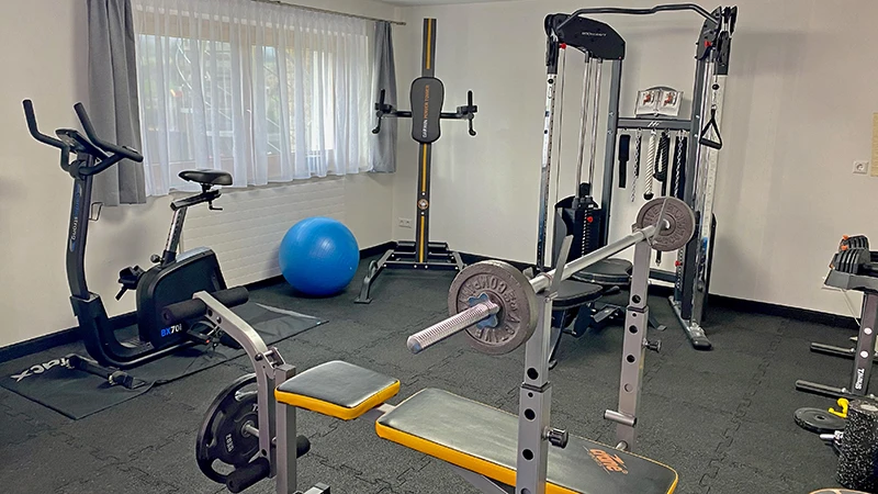 Das Bild zeigt ein gut ausgestattetes Homegym, in dem der Boden mit schwarzen WARCO Fitnessbodenplatten ausgelegt ist. Diese Platten sind an den Kanten verzahnt, wodurch sie formschlüssig und dauerhaft miteinander verbunden sind. Der Fitnessboden bietet eine robuste, rutschfeste und stoßdämpfende Oberfläche, die ideal für intensive Trainingseinheiten geeignet ist. Der Raum ist mit einer Vielzahl von Fitnessgeräten ausgestattet, darunter eine Hantelbank mit Langhantel, ein Ergometer, ein Crosstrainer, eine Kraftstation und verschiedene andere Trainingsgeräte. Ein großes Fenster mit weißen Vorhängen lässt viel Tageslicht in den Raum, was eine angenehme Trainingsatmosphäre schafft. Der schwarze Bodenbelag harmoniert gut mit den modernen Geräten und verleiht dem Homegym ein professionelles und funktionales Ambiente.