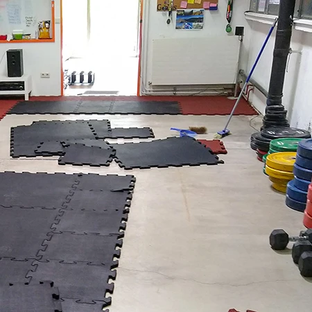 In einem Homegym werden gerade schwarze WARCO Fitnessbodenschutzmatten verlegt. Die Matten bestehen aus robustem Gummigranulat und sind durch die formschlüssige Verzahnung an den Plattenkanten einfach miteinander zu verbinden. Auf der linken Seite des Raumes sind bereits einige Matten ausgelegt, während weitere noch auf den Betonboden gelegt werden. Im Hintergrund sieht man verschiedene Trainingsgeräte wie Hantelscheiben, Kurzhanteln und eine Gewichtsstange. Die Verlegung der Matten schützt den Boden vor Beschädigungen und bietet gleichzeitig eine rutschfeste, stoßdämpfende Oberfläche für intensives Training.