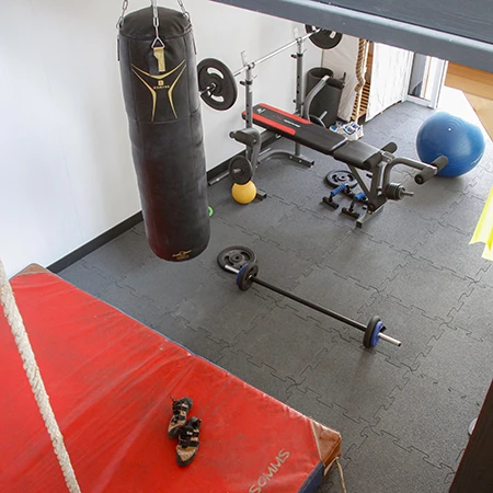 Dieses Homegym ist mit einem schiefergrauen WARCO Fitnessboden ausgestattet, der aus strapazierfähigen Gummigranulatplatten besteht. Die Platten sind durch eine präzise Puzzleverzahnung sicher miteinander verbunden, was eine dauerhafte und formschlüssige Bodenverlegung gewährleistet. Der Raum ist mit einer Vielzahl von Trainingsgeräten ausgestattet, darunter ein Boxsack, eine Hantelbank mit Gewichten, eine Langhantel und ein Gymnastikball. Die robuste und rutschfeste Oberfläche des Bodens bietet hervorragenden Halt und schützt den Untergrund vor Beschädigungen durch schwere Gewichte. Im Vordergrund sieht man eine rote Trainingsmatte, die zusätzlich für dynamische Übungen genutzt wird.