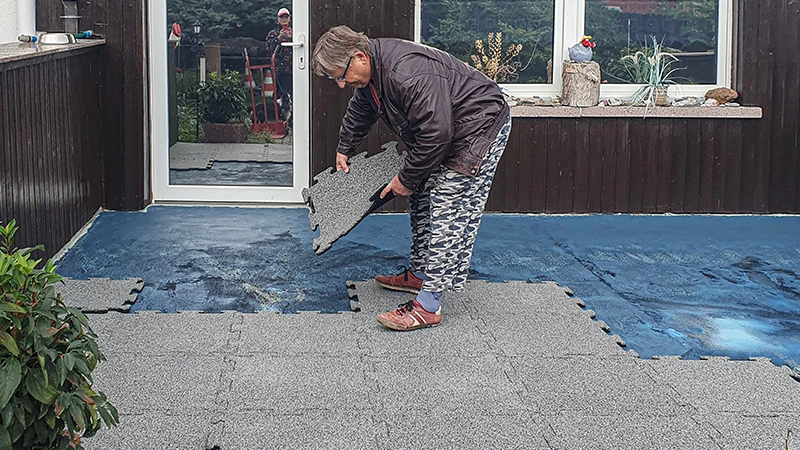 Terasa je v prenovi. Starejša gospa polaga naslednjo 18 mm debelo terasno ploščo s puzzle zaskočnim sistemom na že delno prekrito teraso z novimi ploščami. Tesnilna folija služi kot podlaga za nov gumijasti talni premaz. Natančno spajanje plošč omogoča enostavno in stabilno polaganje.