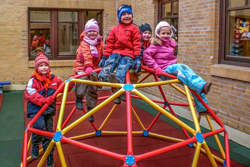 In the inner courtyard of a daycare center stands a spherical climbing frame made of pipes on red and green fall protection panels. A group of children on the climbing frame smiles at the viewer.